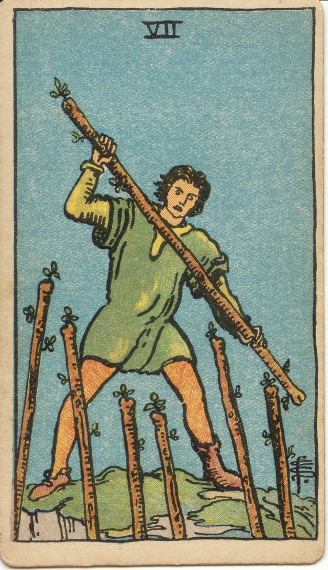 Seven of Wands: The defender on the hill, the six wands from below, the wand held across the body, the mismatched shoes. Rider–Waite–Smith tarot, 1909.